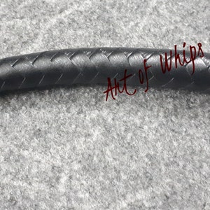 Op de afbeelding: Close-up van een zwarte leren zweep met een gevlochten ontwerp. De zweep is gelabeld "Art of Whips".