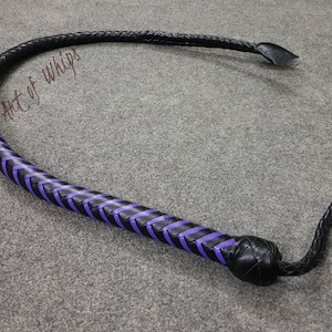 K&ouml;nnte beinhalten: Eine schwarze Lederpeitsche mit violetten geflochtenen Akzenten. Die Peitsche hat einen geflochtenen Griff und einen langen, spitz zulaufenden Schwanz.