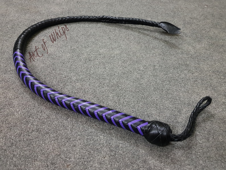 K&ouml;nnte beinhalten: Eine schwarze Lederpeitsche mit violetten geflochtenen Akzenten. Die Peitsche hat einen geflochtenen Griff und einen langen, spitz zulaufenden Schwanz.