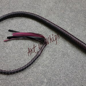 May include: A dark brown leather bullwhip with a braided handle and a burgundy and black tassel. The whip is coiled on a gray surface. The text "Art of Whips" is visible in the image.