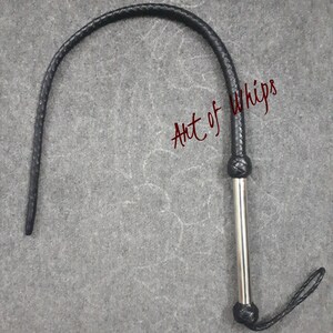 Op de afbeelding: Een zwarte leren zweep met een zilveren handvat en een gevlochten leren lus aan het einde. De zweep is opgerold en het handvat is zichtbaar. De tekst "Art of Whips" is in rood op de achtergrond geschreven.