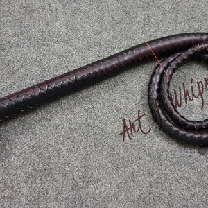 May include: A black and brown leather bullwhip with a braided handle and a long, flowing tail. The whip is coiled and the tail is hanging down. The text "Art Whips" is written on the background.