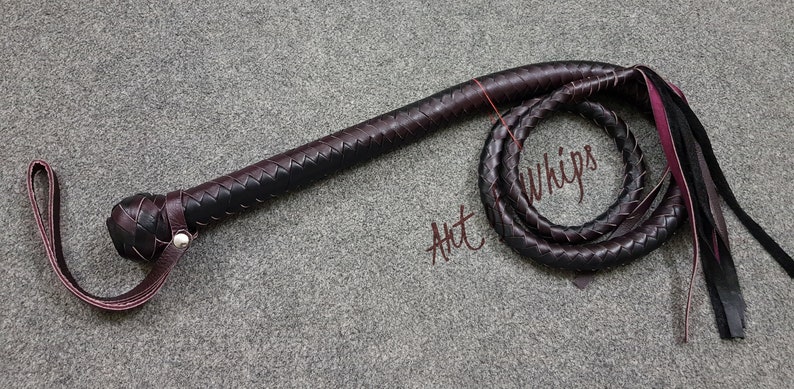 May include: A black and brown leather bullwhip with a braided handle and a long, flowing tail. The whip is coiled and the tail is hanging down. The text "Art Whips" is written on the background.