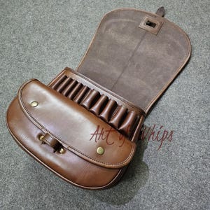 Puede incluir: Un portacartuchos de escopeta de cuero marrón con una solapa que se abre para revelar múltiples portacartuchos. El portacartuchos tiene un cierre de botón de latón y una correa.