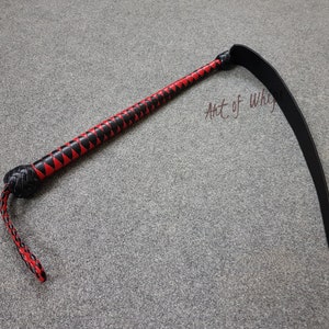 Puede incluir: Un l&aacute;tigo de cuero negro y rojo con un mango trenzado y un l&aacute;tigo largo y curvo. El l&aacute;tigo est&aacute; sobre una alfombra gris. El texto "Art of Whips" es visible en la imagen.