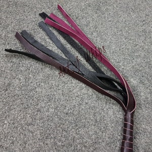 May include: A multi-colored leather whip with a black handle. The whip has black, burgundy, and pink leather strands.