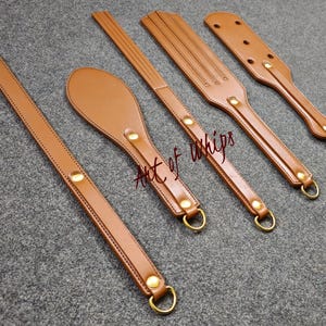 Puede incluir: Cinco látigos de cuero con detalles en latón. Los látigos son marrones y tienen diferentes formas y tamaños. El texto "Art of Whips" es visible en uno de los látigos.
