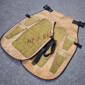 Könnte beinhalten: Eine hellbraune Lederschürze mit mehreren Taschen und schwarzen Riemen. Die Schürze hat ein gestepptes olivgrünes Design auf der Vorderseite und die Worte "Art of Whips" sind auf der Vorderseite aufgedruckt. Die Schürze hat schwarze Schnallen.