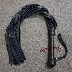 May include: A black leather whip with a braided handle and a long, fringed tail. The whip is coiled and the handle has the text "Art of Whips" embossed on it.