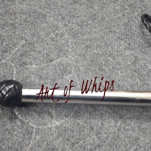 Op de afbeelding: Een zwarte leren zweep met een zilveren metalen kern en een zwarte leren handvat. De zweep is gemarkeerd met "Art of Whips".
