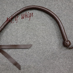 Puede incluir: Un látigo de cuero marrón con un mango trenzado y un extremo anudado. El látigo está enrollado y colocado sobre una superficie gris. El texto "Art of Whips" es visible en la imagen.