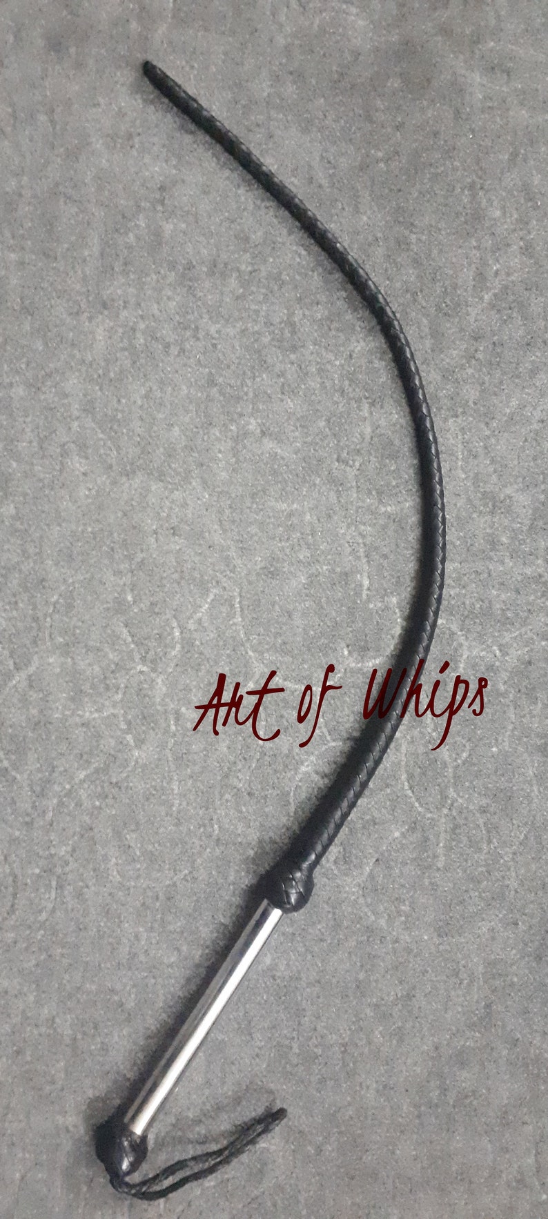 Op de afbeelding: Een zwarte leren zweep met een zilveren handvat. De zweep is opgerold en het handvat is zichtbaar. De tekst "Art of Whips" is zichtbaar op de achtergrond.