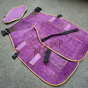 Peut inclure: Chaps en daim violets avec coutures et bordures dorées, avec plusieurs poches et sangles réglables. Comprend un coussinet assorti. Le texte "Art of Whips" est visible sur les chaps.