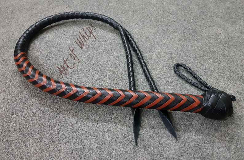 K&ouml;nnte beinhalten: Eine schwarze Lederpeitsche mit einem braunen geflochtenen Lederdesign. Die Peitsche hat einen geflochtenen Griff und einen langen, d&uuml;nnen Schwanz. Der Text "Art of Whips" ist auf der Peitsche sichtbar.