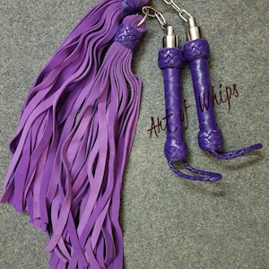 May include: Two purple leather whips with braided handles and tassels. The handles are connected by a silver chain. The text "Art of Whips" is visible in the image.