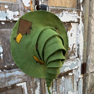 Puede incluir: Un sombrero de fieltro verde con una copa alta en forma de cono en espiral y un ala ancha. El sombrero presenta parches rectangulares decorativos de color marrón y amarillo cosidos en el ala con hilo negro visible y texturizado.
