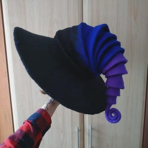 Op de afbeelding: Een zwarte heksenhoed met een lange, puntige rand en een spiraalvormige, gelaagde staart in tinten blauw en paars. De hoed is gemaakt van een zacht, viltachtig materiaal.