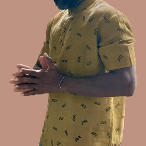 May include: Mustard yellow short-sleeved shirt with a mandarin collar and a pattern of black wave-like shapes. The shirt has a button-down front and rolled-up sleeves. The shirt is made of a lightweight fabric.