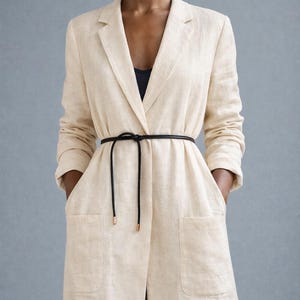 May include: A long, cream-colored blazer with a black belt cinching the waist. The blazer has two large front pockets and a notched lapel. The belt is a thin, black cord with gold-colored metal tips. The blazer is made of a textured fabric.