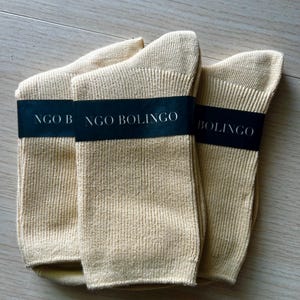 Puede incluir: Tres pares de calcetines beige est&aacute;n cuidadosamente dispuestos sobre una superficie de madera clara. Cada par est&aacute; envuelto con una banda negra que dice "NGO BOLINGO" en texto blanco. Los calcetines parecen estar hechos de tejido de punto acanalado.