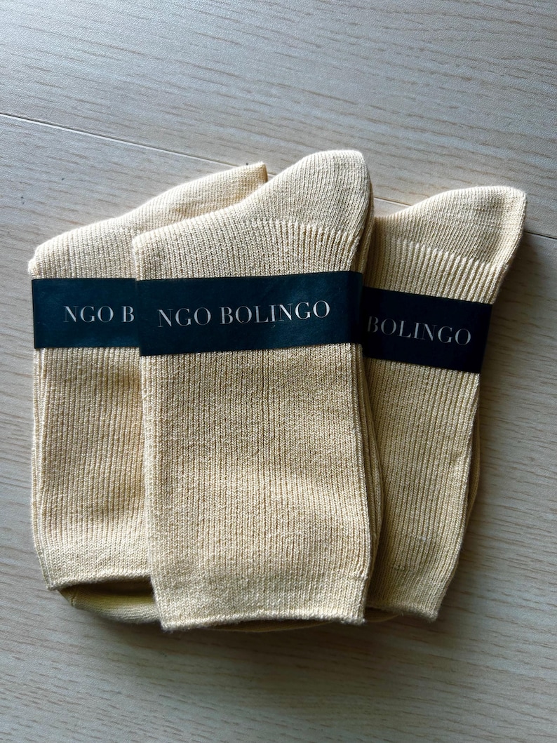 Puede incluir: Tres pares de calcetines beige est&aacute;n cuidadosamente dispuestos sobre una superficie de madera clara. Cada par est&aacute; envuelto con una banda negra que dice "NGO BOLINGO" en texto blanco. Los calcetines parecen estar hechos de tejido de punto acanalado.