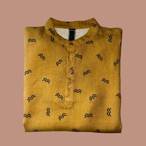 May include: Mustard-colored shirt with a mandarin collar and a button-down placket. The shirt is patterned with small, black, wave-like designs. The shirt is folded neatly and is made of a textured fabric.