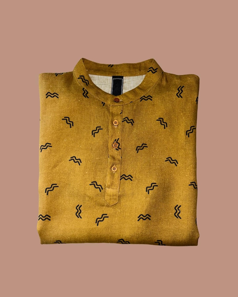 May include: Mustard-colored shirt with a mandarin collar and a button-down placket. The shirt is patterned with small, black, wave-like designs. The shirt is folded neatly and is made of a textured fabric.