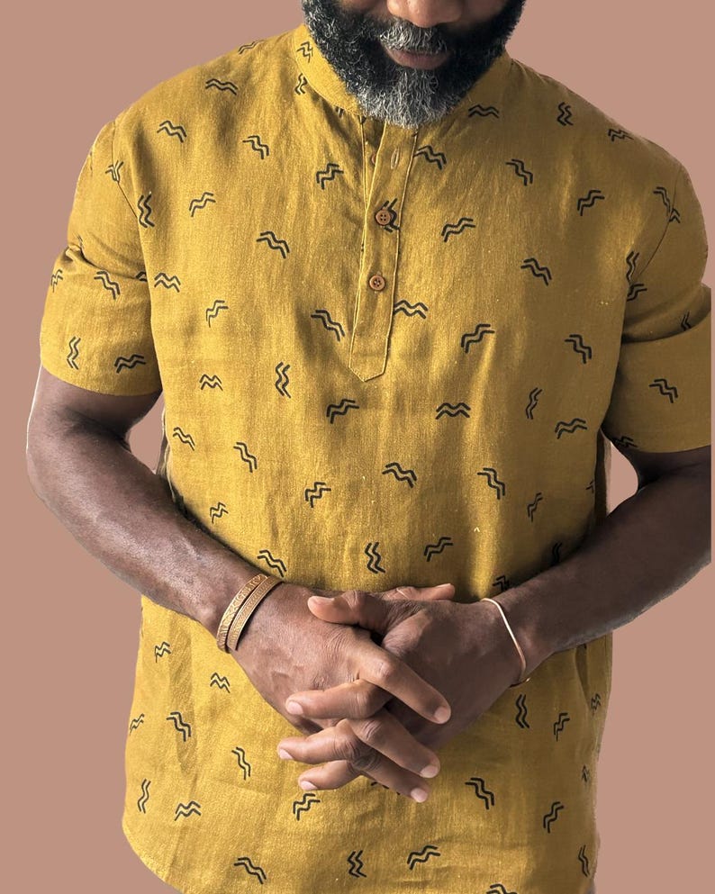May include: Mustard yellow short-sleeved shirt with a mandarin collar and a small buttoned placket. The shirt is patterned with small, black, wave-like designs. The person is wearing a gold bracelet and a silver bracelet.