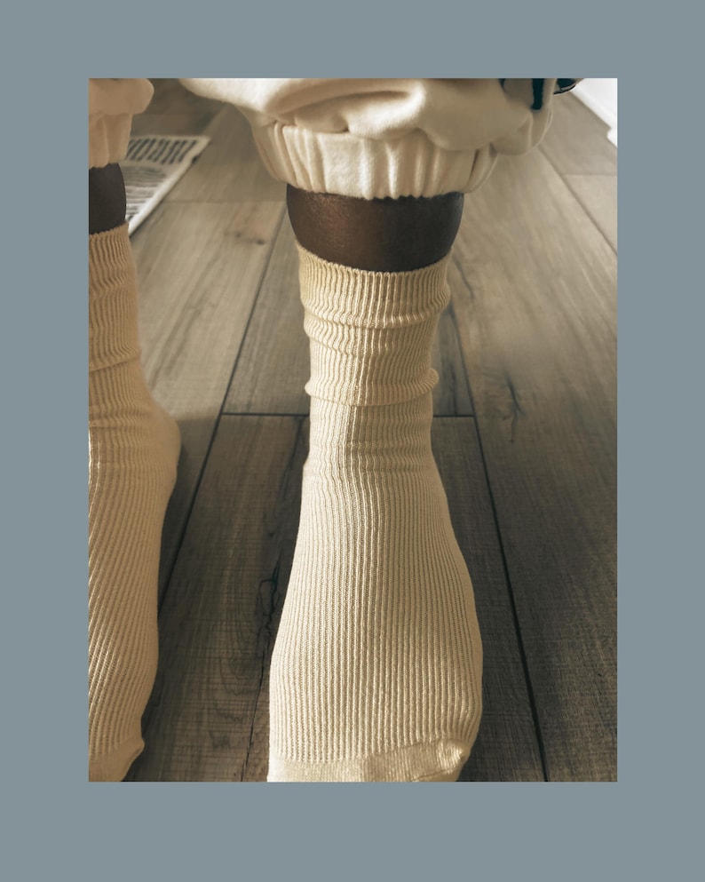 Puede incluir: Primer plano de calcetines acanalados de color crema. Los calcetines est&aacute;n subidos, mostrando la parte inferior de la pierna y el tobillo. Los calcetines est&aacute;n hechos de un material suave y texturizado. El fondo es un suelo de madera marr&oacute;n claro.