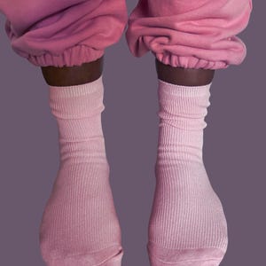 Puede incluir: Se ven dos pares de calcetines de canal&eacute; rosa claro, que se llevan con un pantal&oacute;n de ch&aacute;ndal rosa a juego. Los calcetines est&aacute;n subidos, con la tela recogida alrededor de los tobillos. El fondo es morado.