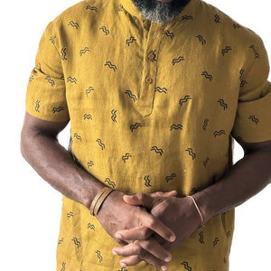 May include: Mustard yellow short-sleeved shirt with a mandarin collar and a small, repeating black wave pattern. The shirt has a buttoned placket and is worn with a brown bracelet and a copper bracelet.
