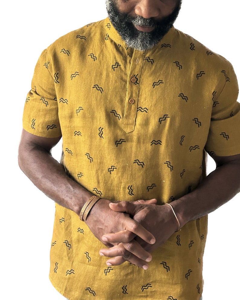 May include: Mustard yellow short-sleeved shirt with a mandarin collar and a small, repeating black wave pattern. The shirt has a buttoned placket and is worn with a brown bracelet and a copper bracelet.