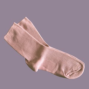 Puede incluir: Un par de calcetines rosa p&aacute;lido, hechos de tejido de punto acanalado. Los calcetines est&aacute;n doblados, mostrando el tal&oacute;n y los dedos. El fondo es morado apagado.
