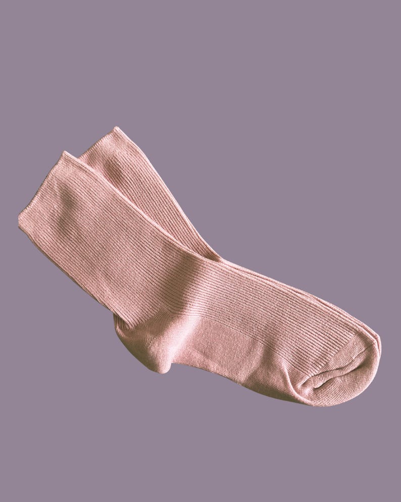 Puede incluir: Un par de calcetines rosa p&aacute;lido, hechos de tejido de punto acanalado. Los calcetines est&aacute;n doblados, mostrando el tal&oacute;n y los dedos. El fondo es morado apagado.