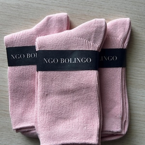 Puede incluir: Tres pares de calcetines rosa claro apilados. Cada par est&aacute; envuelto con una banda negra que dice "NGO BOLINGO" en texto blanco. Los calcetines est&aacute;n hechos de tejido de punto acanalado.