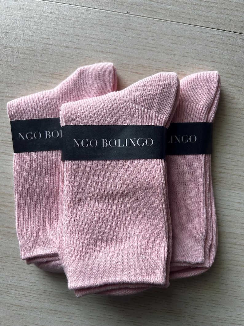 Puede incluir: Tres pares de calcetines rosa claro apilados. Cada par est&aacute; envuelto con una banda negra que dice "NGO BOLINGO" en texto blanco. Los calcetines est&aacute;n hechos de tejido de punto acanalado.