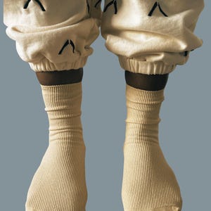 Puede incluir: Primer plano de calcetines acanalados de color crema y pantalones de ch&aacute;ndal a juego. Los pantalones de ch&aacute;ndal tienen dise&ntilde;os de chevr&oacute;n negros cerca de los tobillos. Los calcetines est&aacute;n subidos y los pantalones est&aacute;n recogidos en los tobillos. El fondo es azul liso y apagado.