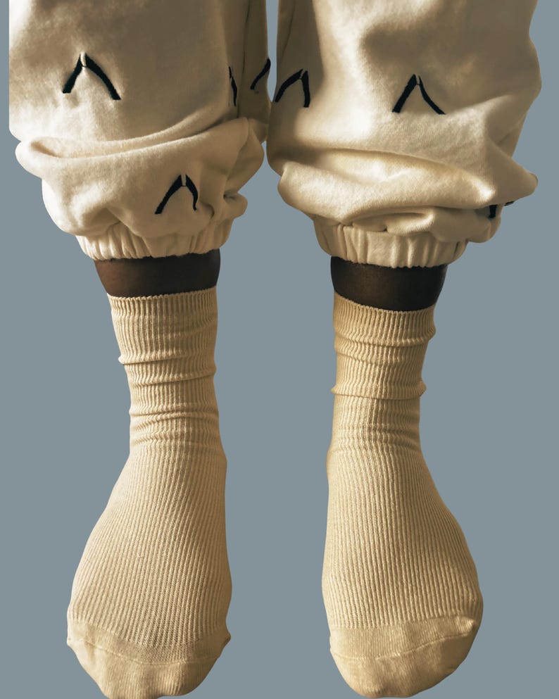 Puede incluir: Primer plano de calcetines acanalados de color crema y pantalones de ch&aacute;ndal a juego. Los pantalones de ch&aacute;ndal tienen dise&ntilde;os de chevr&oacute;n negros cerca de los tobillos. Los calcetines est&aacute;n subidos y los pantalones est&aacute;n recogidos en los tobillos. El fondo es azul liso y apagado.