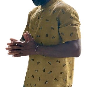 May include: Mustard-colored short-sleeved shirt with a mandarin collar and a pattern of black wave-like shapes. The shirt has a button-down front and is made of a lightweight fabric. The shirt is part of a casual clothing collection.
