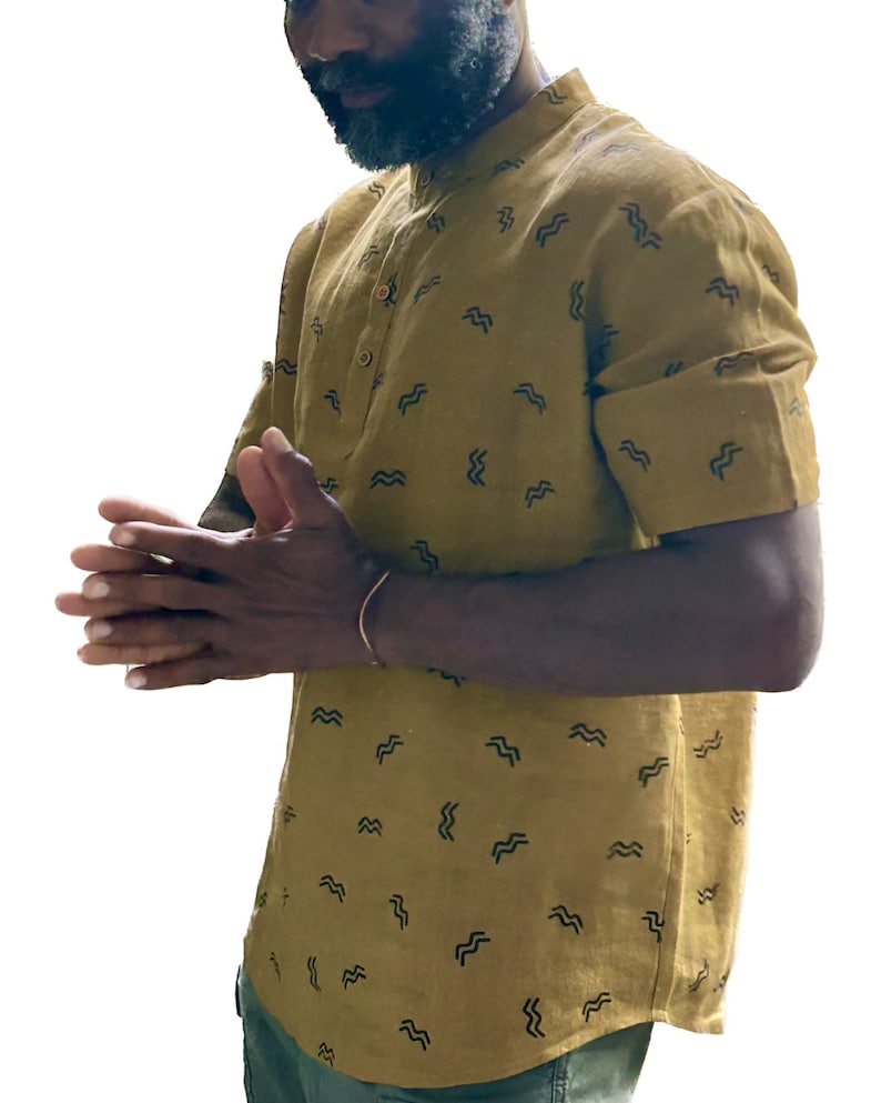 May include: Mustard-colored short-sleeved shirt with a mandarin collar and a pattern of black wave-like shapes. The shirt has a button-down front and is made of a lightweight fabric. The shirt is part of a casual clothing collection.