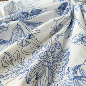 Pode incluir: Close-up de tecido branco com bordado floral azul detalhado. O design apresenta contornos detalhados de flores e folhas, criando um padrão texturizado e elegante. O tecido está dobrado, mostrando a profundidade e dimensão do design.
