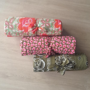 May include: Three rolled quilted blankets tied with fabric strips. The top blanket features a bright orange and green floral pattern. The middle blanket has a pink and brown leopard print. The bottom blanket displays an olive green paisley design.