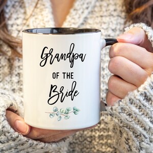 Może przedstawiać: Biały kubek ceramiczny z czarną obw&oacute;dką i uchwytem. Kubek ma czarny napis "Grandpa of the Bride" z małym zielonym wzorem kwiatowym na dole.