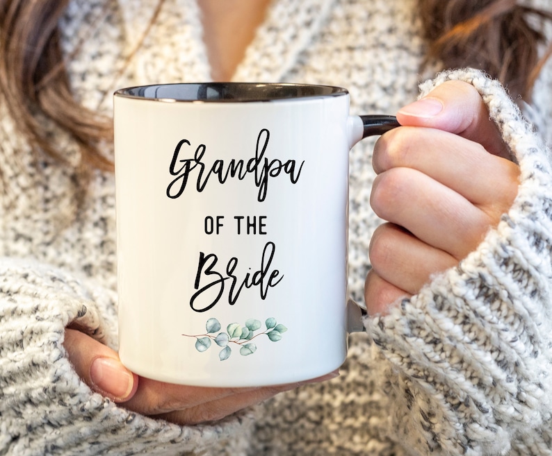 Może przedstawiać: Biały kubek ceramiczny z czarną obw&oacute;dką i uchwytem. Kubek ma czarny napis "Grandpa of the Bride" z małym zielonym wzorem kwiatowym na dole.