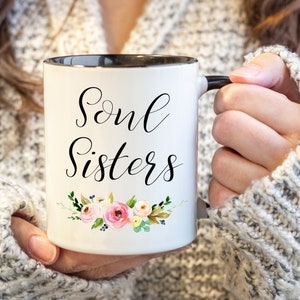 May include: White ceramic mug with black rim and handle, featuring the words "Soul Sisters" in elegant script. A floral design with pink and yellow roses, green leaves, and blue accents decorates the bottom of the mug.
