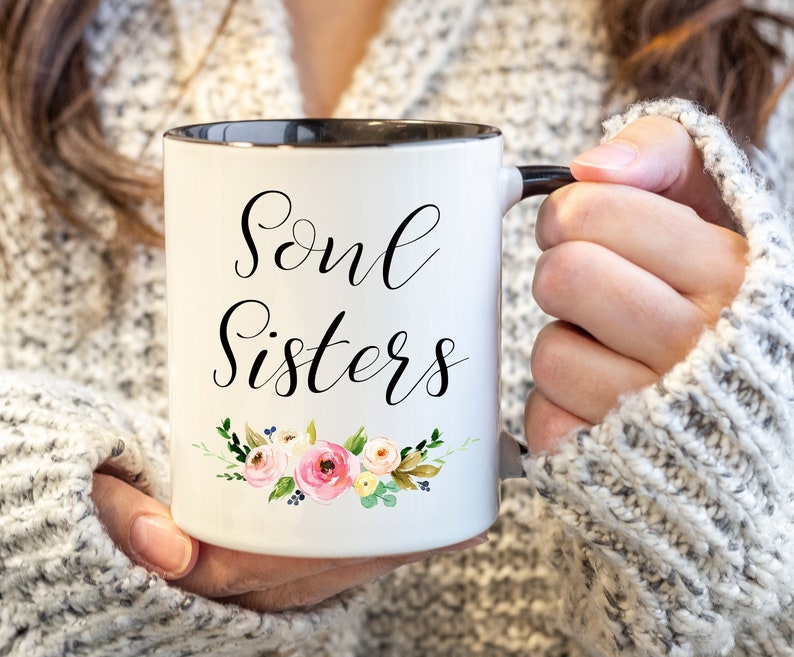 May include: White ceramic mug with black rim and handle, featuring the words "Soul Sisters" in elegant script. A floral design with pink and yellow roses, green leaves, and blue accents decorates the bottom of the mug.