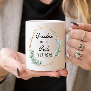 Pode incluir: Caneca de cer&acirc;mica branca com moldura geom&eacute;trica dourada e folhas de eucalipto verdes. O texto diz "Grandma of the Bride 10.17.2020".