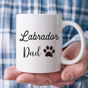 Puede incluir: Taza de cer&aacute;mica blanca con las palabras "Labrador Dad" en escritura negra y una huella de pata negra. La taza tiene un asa curvada.