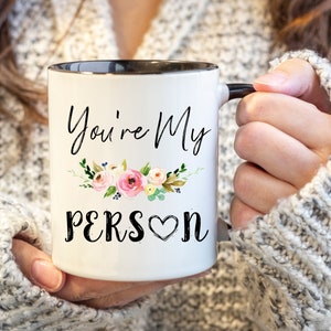 K&ouml;nnte beinhalten: Eine wei&szlig;e Keramiktasse mit schwarzem Rand und Henkel. Die Tasse hat ein Blumenmuster und den Text "You're My Person" mit einem Herzen.
