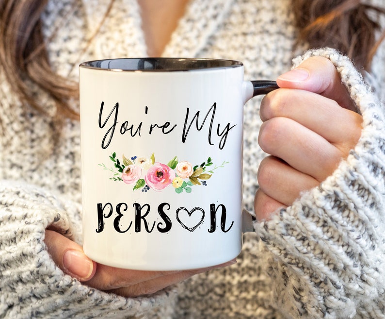 K&ouml;nnte beinhalten: Eine wei&szlig;e Keramiktasse mit schwarzem Rand und Henkel. Die Tasse hat ein Blumenmuster und den Text "You're My Person" mit einem Herzen.
